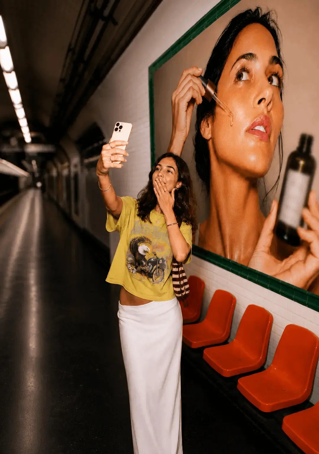 Une jeune influenceuse se prend en photo avec sa campagne publicitaire affichée derrière elle, dans le métro parisien